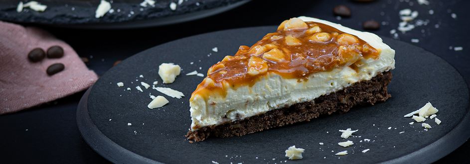 Ein Stück White Chocolate Cheesecake Mit Macadamia-Karamell-Topping auf einem dunklen Teller