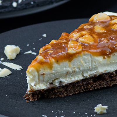 Ein Stück White Chocolate Cheesecake Mit Macadamia-Karamell-Topping auf einem dunklen Teller Ein Stück White Chocolate Cheesecake Mit Macadamia-Karamell-Topping auf einem dunklen Teller
