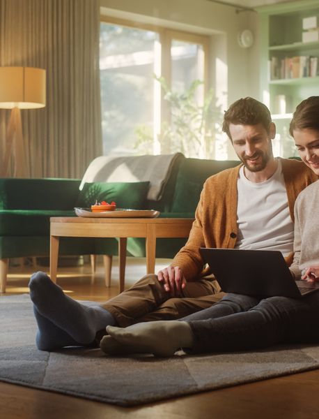Pärchen sitzt auf dem Boden, vor dem Sofa und schaut auf Laptop
