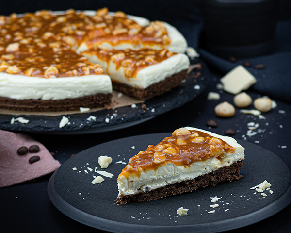Ein Stück White Chocolate Cheesecake Mit Macadamia-Karamell-Topping angerichtet neben Kuchen